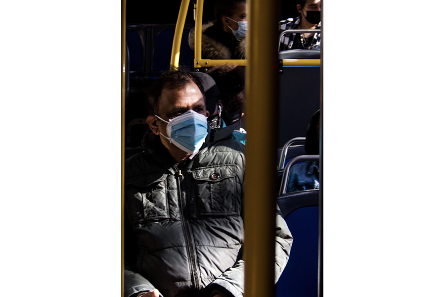Three people wearing face coverings seen through the yellow support poles throughout the bus. 