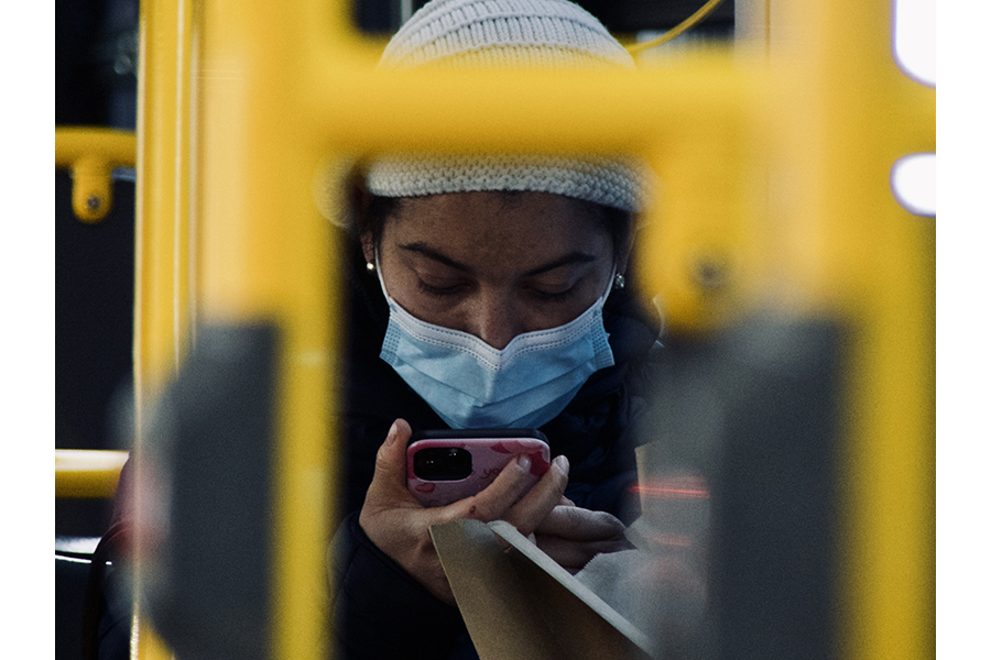 Portrait a young woman, seated on Manhattan bus, wearing a beanie and face covering, staring intently at her phone. 