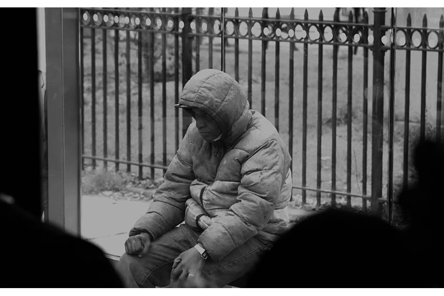 A man as seen from the inside of the bus, in a hooded winter coat, sitting in a daze at a Harlem bus stop. 