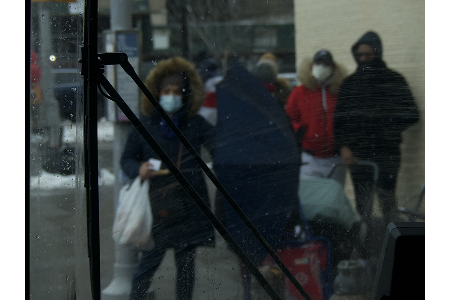 : Through the front windshield of the bus, a woman is seen holding out an umbrella. Behind her stands people with luggage, waiting for the local Bronx bus. 