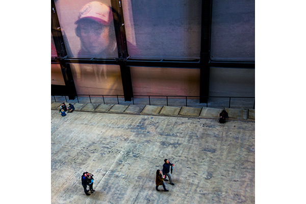 View of a sidewalk with a few pedestrians from a second-story window, the faint reflection of the photographer in view. 