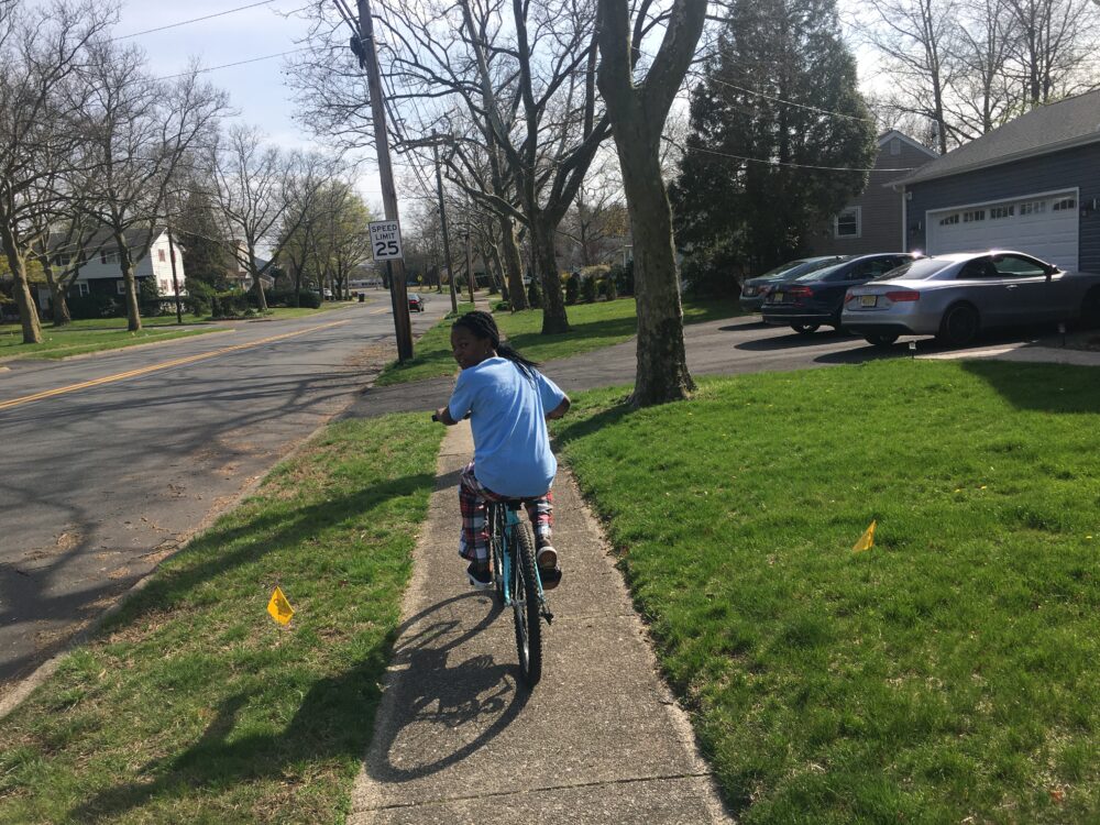 A girl riding a bicycle looks back over her shoulder; she rides down a suburban sidewalk, flanked by grass on both sides.