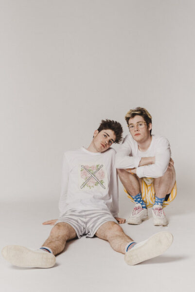 The Burns brothers shown together on a white studio background, wearing complementary striped shorts and long-sleeve T-shirts emblazoned with their instruments; on the left Edie sits and Iz crouches, and on the right Iz has his arm around Eddie.