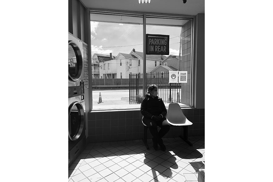 A woman sits in a laundromat. She has a mask on and is sitting in front of a window; black and white.