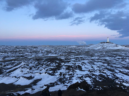 A photograph of a snowy-covered rocky expanse, under a light blue sky, with pink at the horizon.. Lighthouse in the distance. 