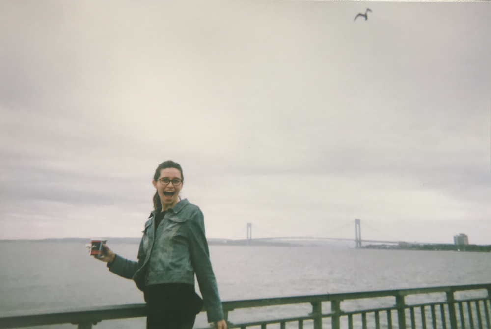 Hannah smiles wide, bakery coffee in hand, along the South Brooklyn waterfront.
