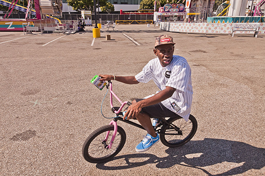 Tyler, the Creator, wearing a snap-back cap on a bicycle in an empty parking lot. 