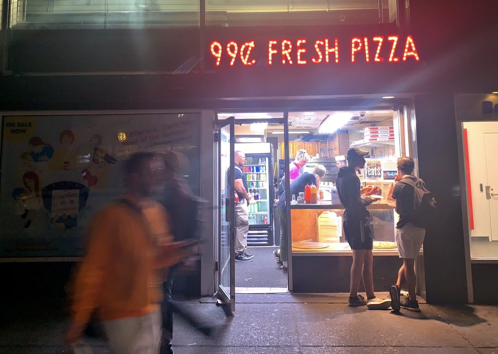 Photograph of a dollar pizza storefront at night, neon sign reading "99-cent Fresh Pizza" illuminated