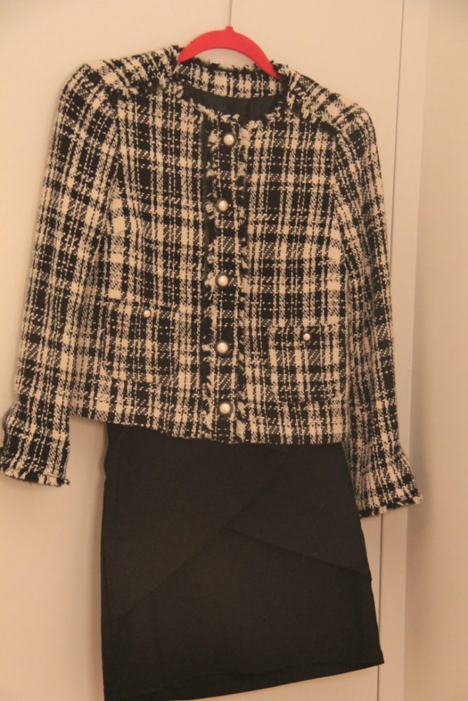 An outfit on a hanger: a plaid jacket with white buttons, pearls, and a black skirt. 