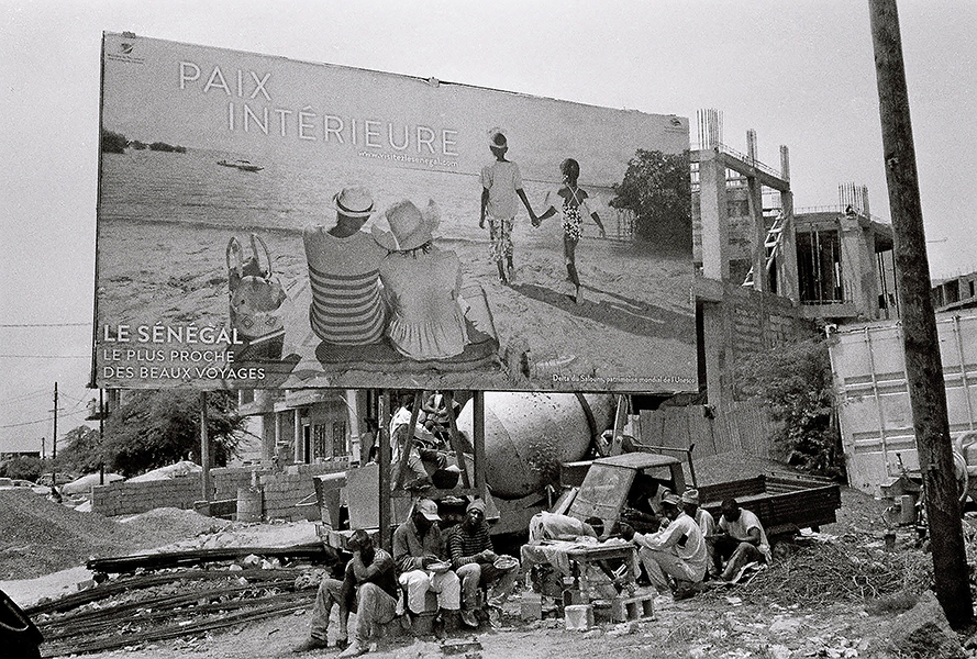b&w photo of billboard depicting vacation inSenegal, workers eat below