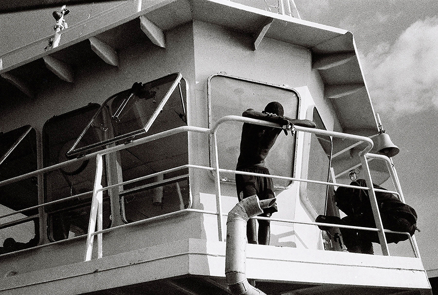 A man leans against the railing of a boat deck, his head between his arms. 