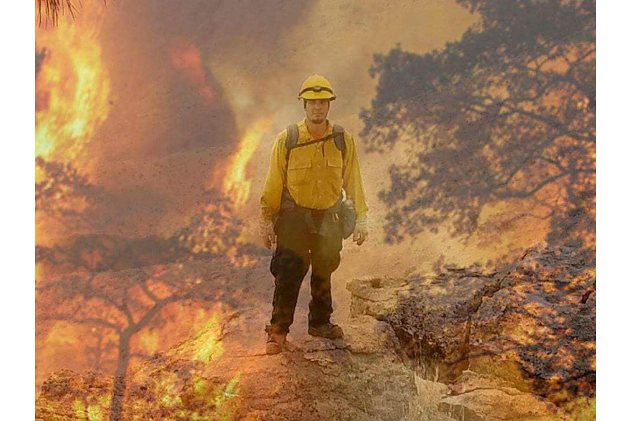 Photograph of Jimmy in firefighting gear as a fire blazes around him; burning tree branches are visible in the background
