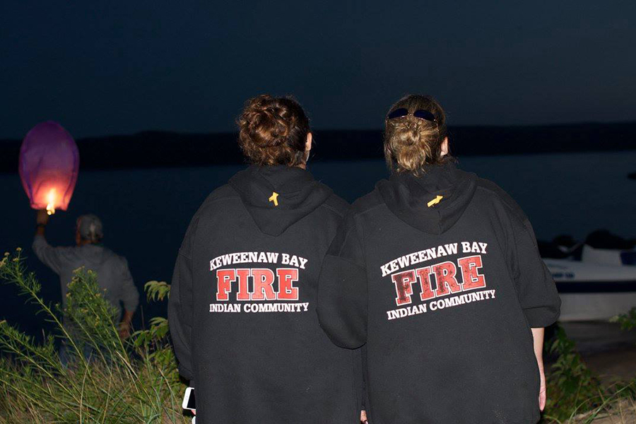 Two women, seen from the back wearing Keweenaw Bay Indian Community Fire Department sweatshirts, seated by a lake at night.