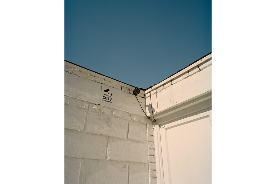 Photograph where two white concrete walls come to a corner; a security camera is near the center of the frame, blue sky in the background