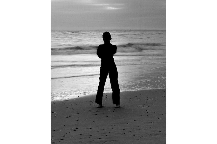 Black-and-white photograph of a person at the edge of the sea, back turned, in silhouette