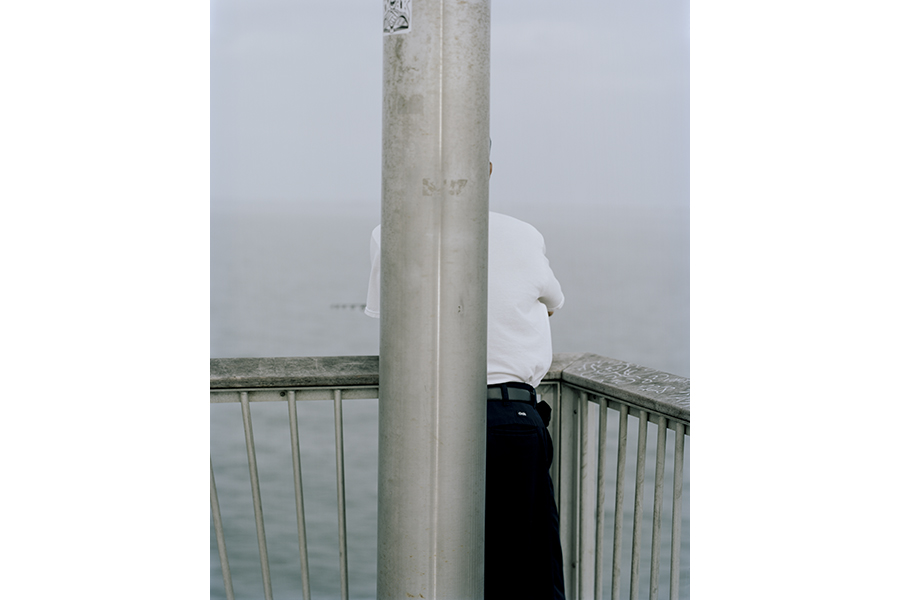 Photograph of a man standing at a railing overlooking the sea; a large pole in the foreground obscures most of his form
