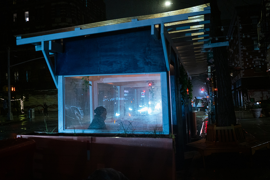  Shadowy figure sits alone at a nondescript foodshed; car lights reflect off the shed's plastic windows.