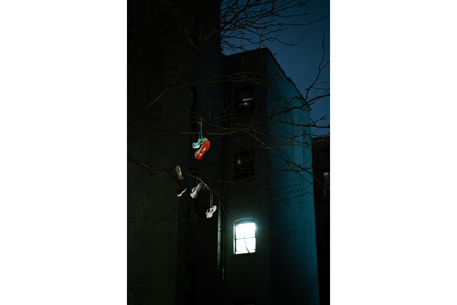 Pairs of shoes hanging on power lines above a single lit window. 