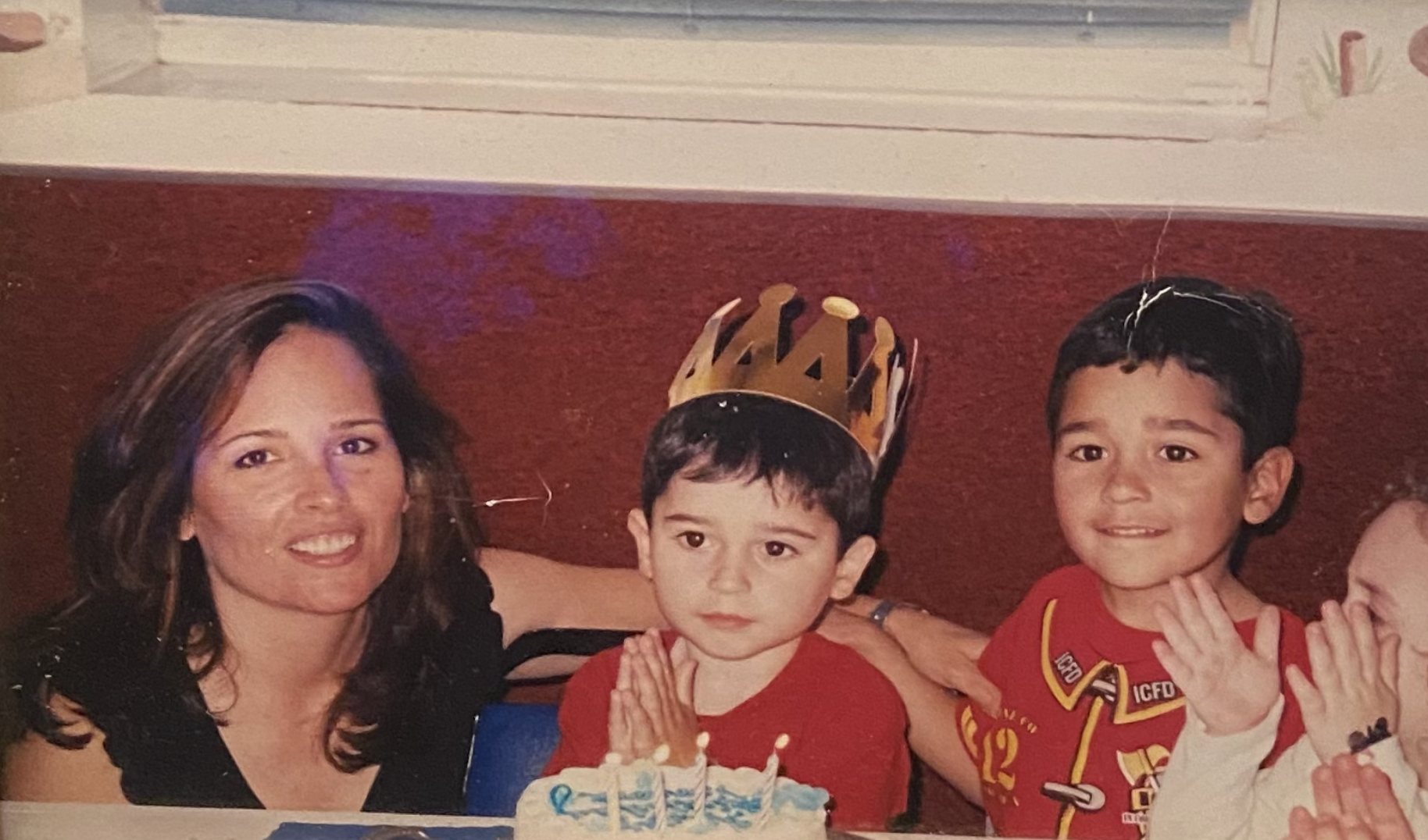 A mother sits with her two young boys, the boy at center wearing a birthday a paper crown and a lit birthday cake in front of him, and the boy at right wearing a fire-fighter costume