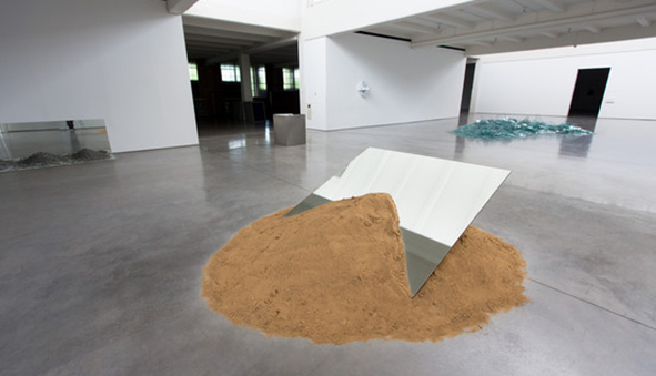 Sculpture of a tilted mirror resting in a pile of sand.