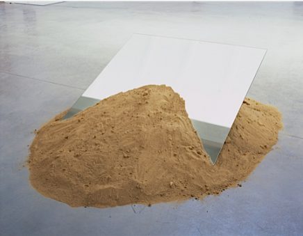 Sculpture of a tilted mirror resting in a pile of sand.
