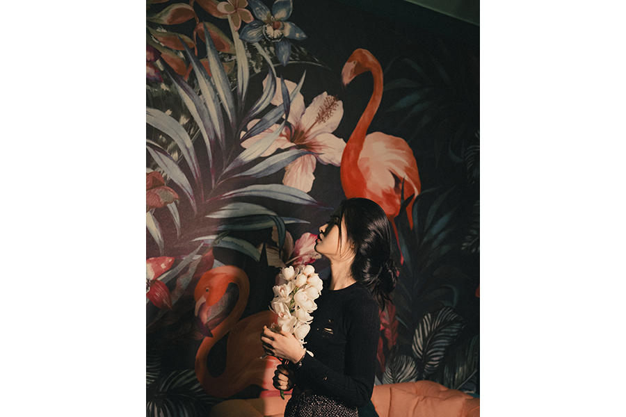 A woman, in profile, holds a spray of white orchids; she stands against a dark wallpaper with a tropical pattern, flamingos, hibiscus, and palm fronds are all to be seen