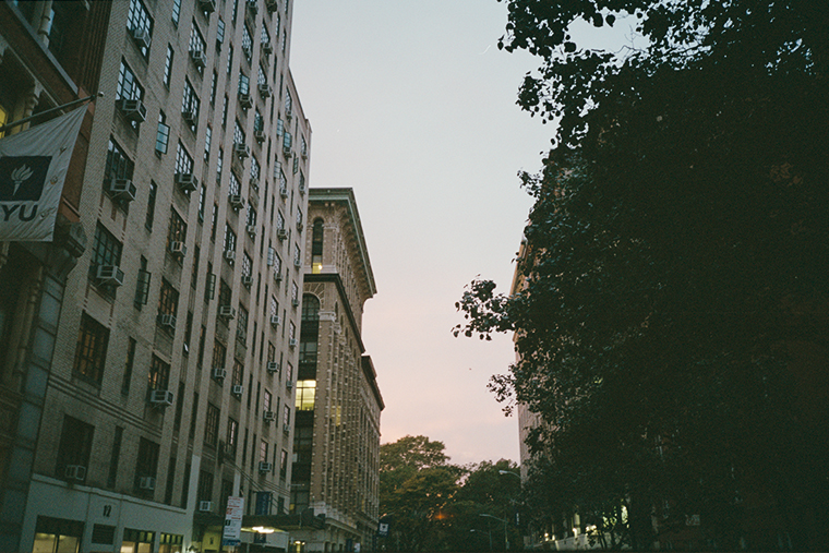 Photograph of a street, angled upwards to show gray-blue and pink sky; high NYU buildings to the left, and trees to the right. Stoplight and park in the distance.
