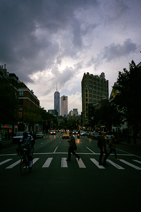 Photograph at an intersection under a cloudy sky, pedestrians crossing, oncoming traffic in background, One World Trade Center in distance.