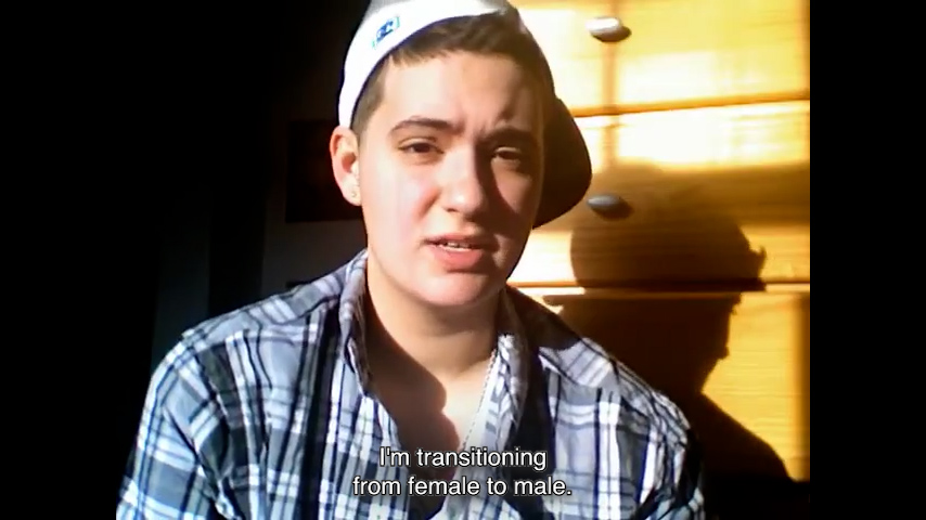 Webcam still image of a boy facing the camera. Subtitles: “I’m transitioning from female to male.”
