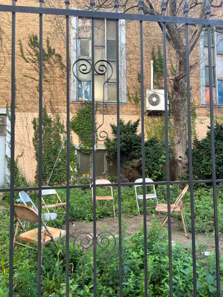 Six metal chairs behind a black gate arranged in a half circle in a neglected garden front of an old building ornamented with ivy.