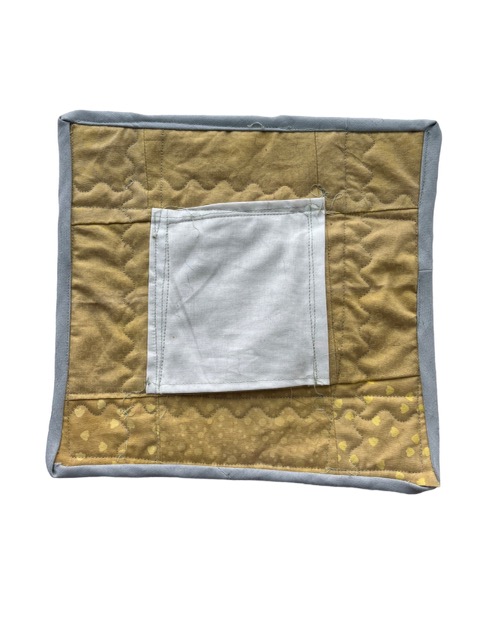 The back of the quilt has a pocket in the center of it made with a white fabric. Behind and underneath the pocket is a yellow fabric, which is dyed with turmeric. The yellow fabric makes up the entirety of the back of the quilt, and has varying patterns on it, including dots, florals, and leaves.