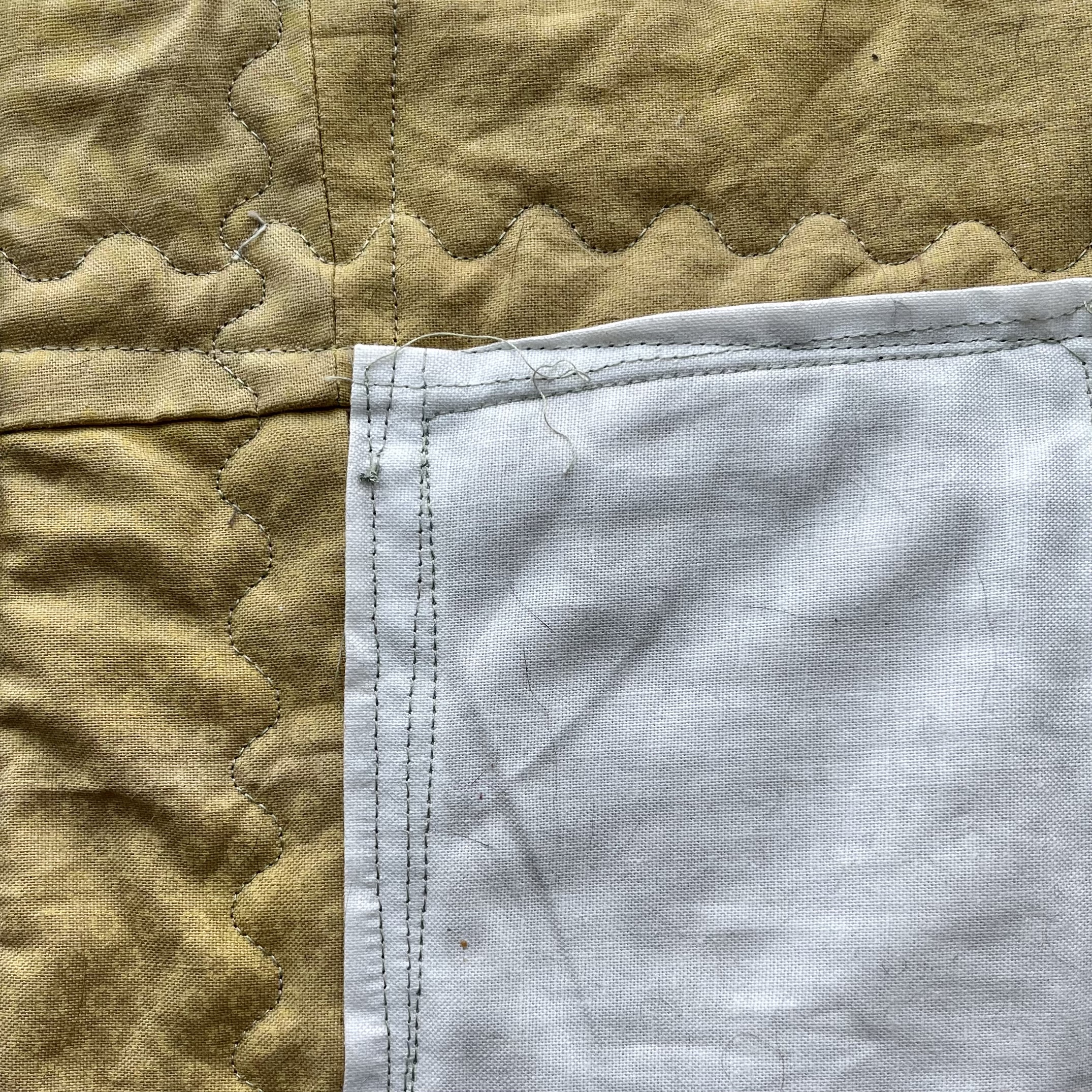 This image shows a closer look at the turmeric-dyed fabric, white pocket, and stitching. The stitching is a light green color. The pocket is stitched on with a straight stitch. The quilt top, batting, and backing are stitched together using a wave pattern. 