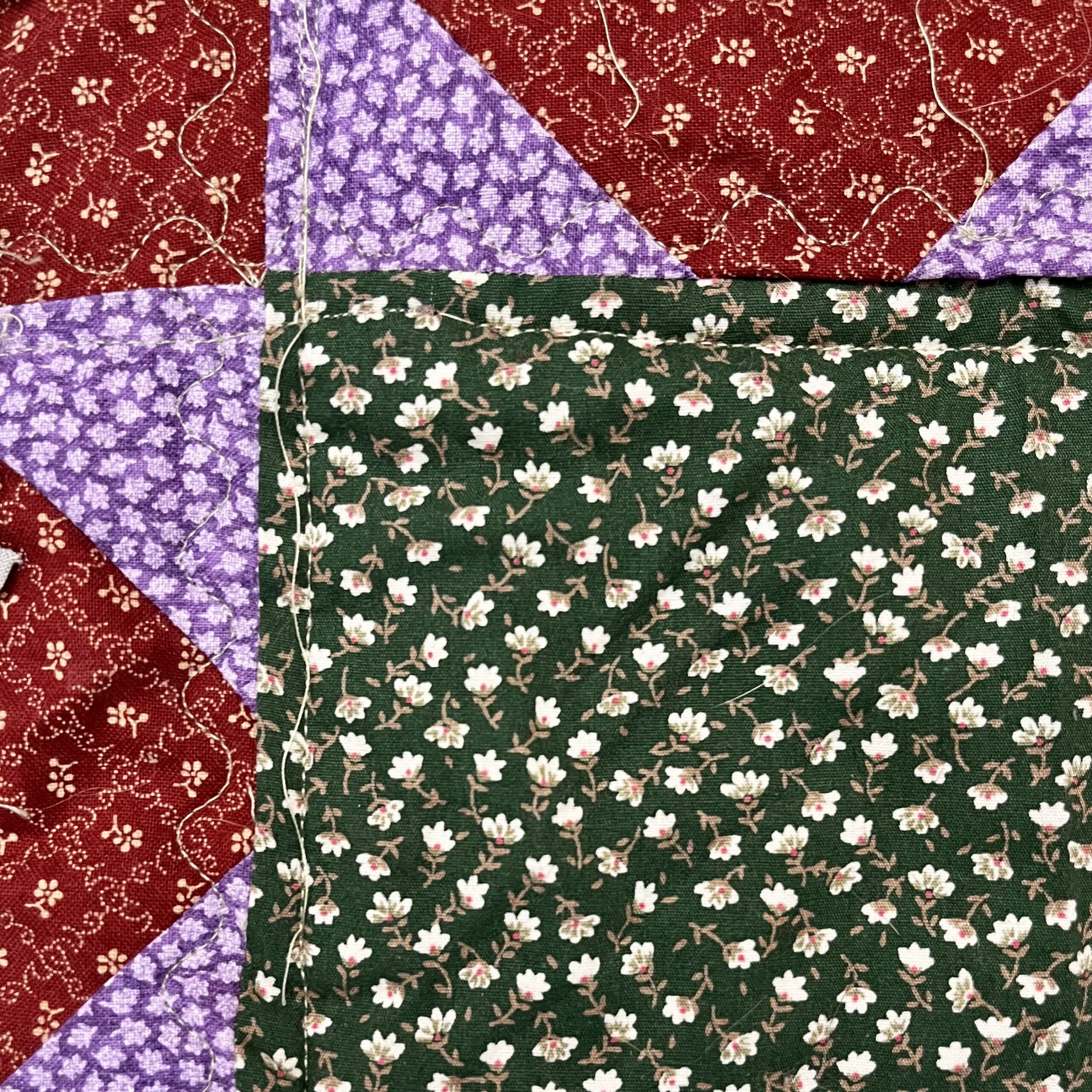 This image shows a closer look at one corner of the sawtooth star pattern on the front of the quilt. One corner of the green square with a floral pattern is visible. Four purple floral triangles are visible, as well as three red squares. There is also visible stitching in green thread. 