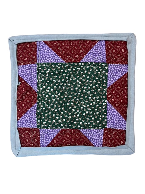 This is an image of the front of the quilt. The quilt top is made in a sawtooth star pattern. At the center of the star is a square of dark green fabric with a floral pattern of repeated white and light brown flowers and light green leaves. There are eight triangles emerging from the square at the center, two on each side of the square. The triangles are a lavender fabric with a pattern of small white flowers on them. Between each triangle is a square of red fabric. The red fabric has a squiggly diamond pattern on it. In the center of each diamond is a small white flower. The quilt is stitched with a light green thread. The quilt is bound with a grey fabric.