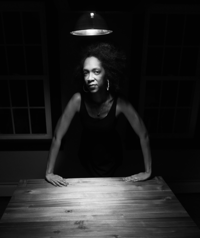 Black-and-white portrait of Shatima Jones standing at the head of a dining table, hands firmly pressing into the wood and gazing into the camera, spotlight by a lamp hanging above