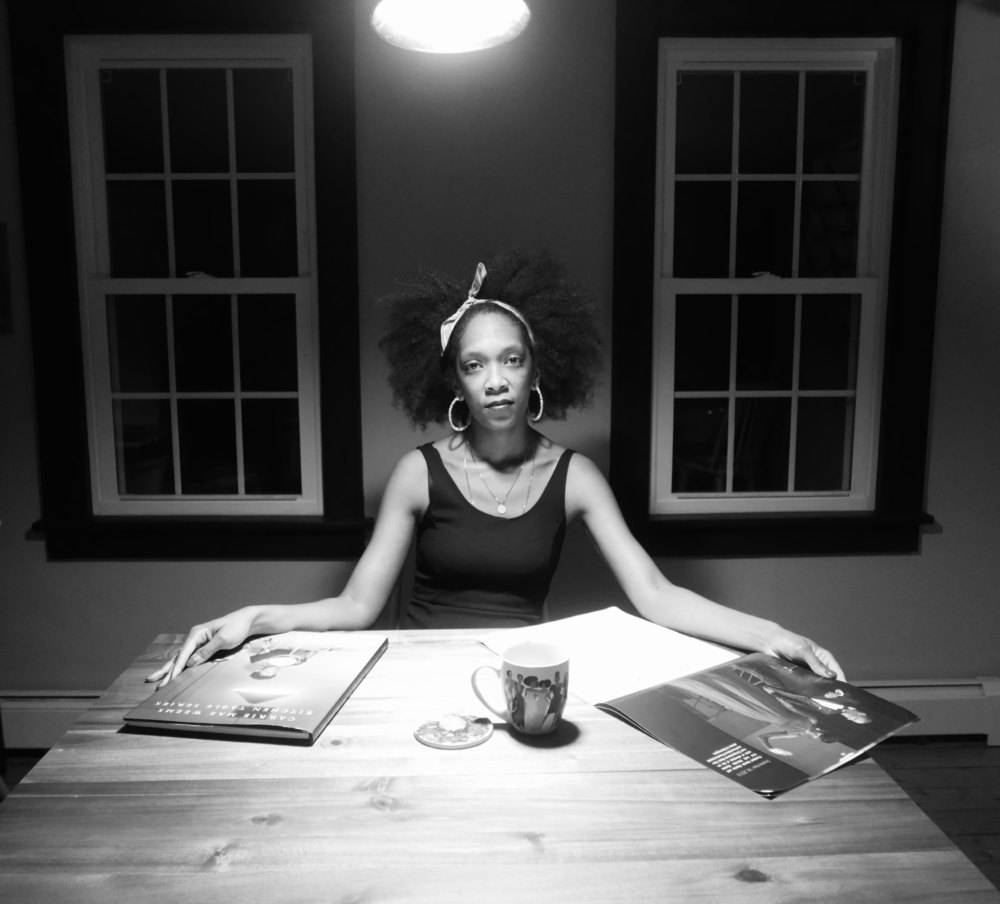 Black-and-white portrait of Shatima Jones seated at the head of a table, gazing directly into the camera, hair tied with a bandana; on the table are a book of Carrie Mae Weem's photography, a coffee mug, and an open calendar.